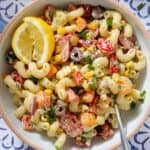 Colorful creamy pasta salad with diced veggies and two lemon wedges in a low bowl with a beige rim and with a fork on the right side.