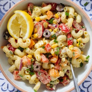 Colorful creamy pasta salad with diced veggies and two lemon wedges in a low bowl with a beige rim and with a fork on the right side.