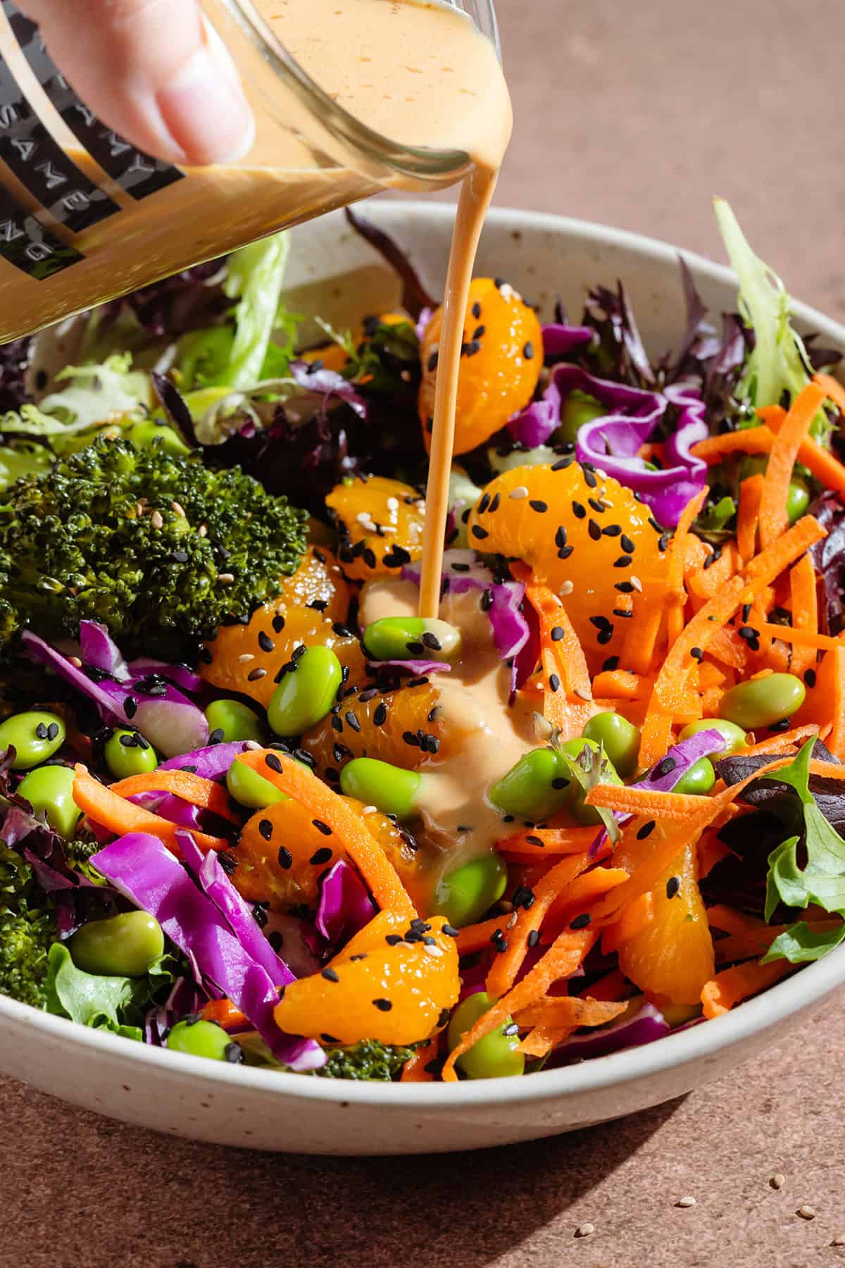 Creamy dressing being poured over a salad with edamame, shredded carrots, cabbage, and tangerine pieces.