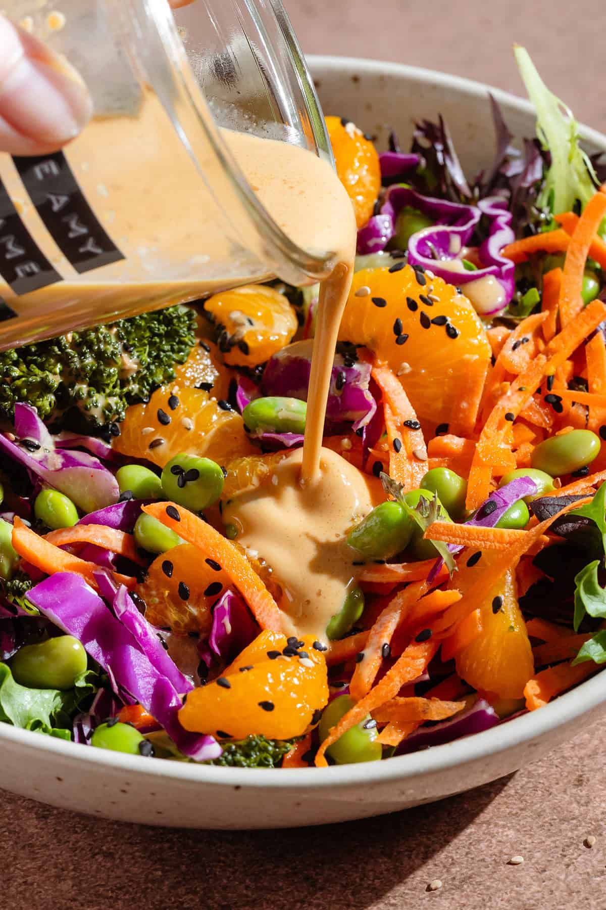 Creamy dressing being poured over a salad with edamame, shredded carrots, cabbage, and tangerine pieces.