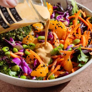 Creamy dressing being poured over a salad with edamame, shredded carrots, cabbage, and tangerine pieces.