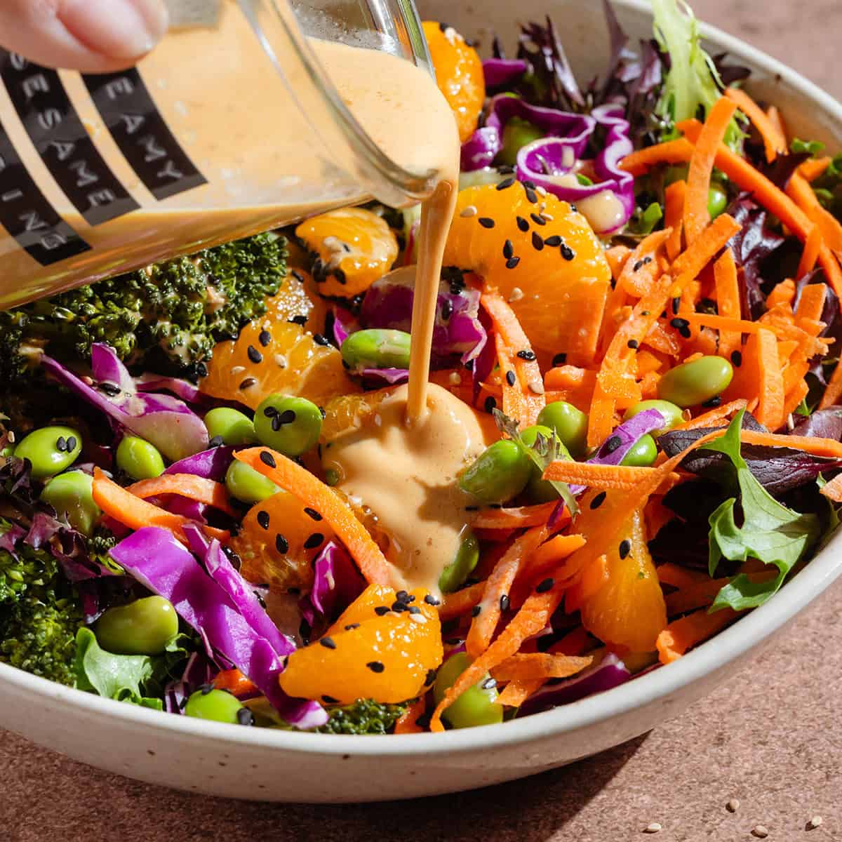 Creamy dressing being poured over a salad with edamame, shredded carrots, cabbage, and tangerine pieces.