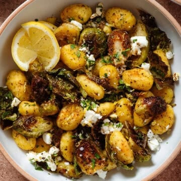 Crispy golden roasted brussels sprouts with gnocchi and feta in a white bowl garnished with lemon zest and two lemon wedges on the left.