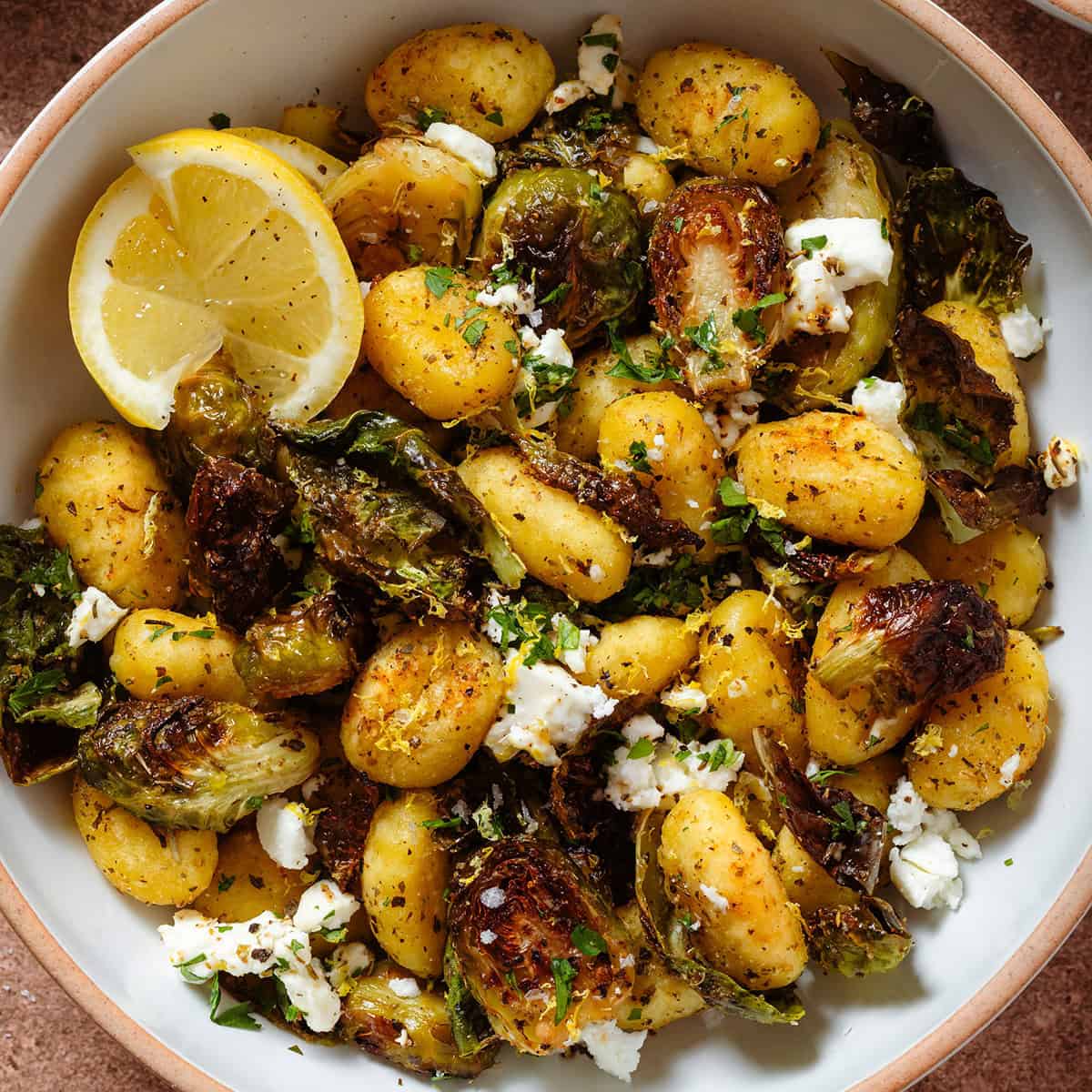 Crispy golden roasted brussels sprouts with gnocchi and feta in a white bowl garnished with lemon zest and two lemon wedges on the left.