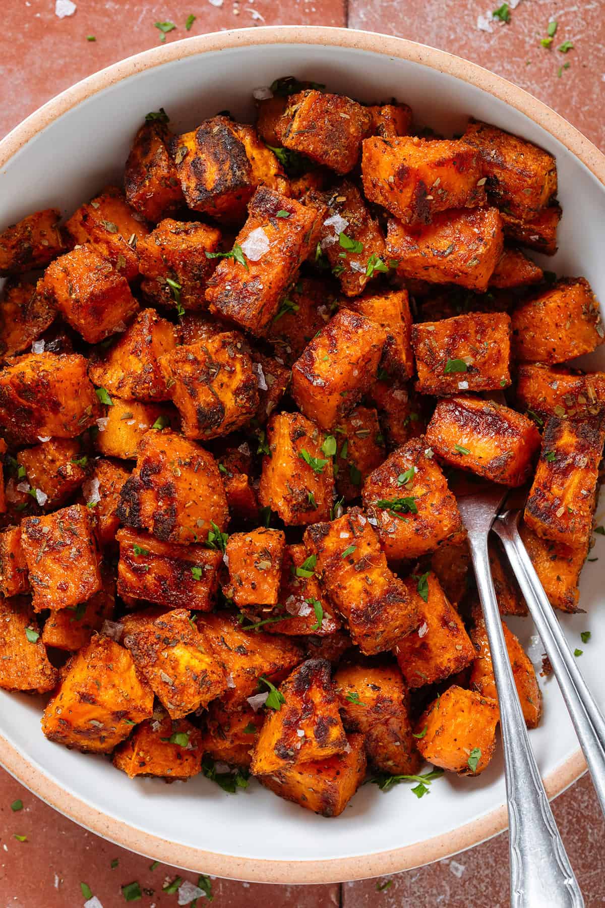 Crispy charred roasted diced sweet potatoes in a white bowl with a beige rim sprinkled with flaky sea salt and finely chopped fresh parsley and with two serving forks on the right.