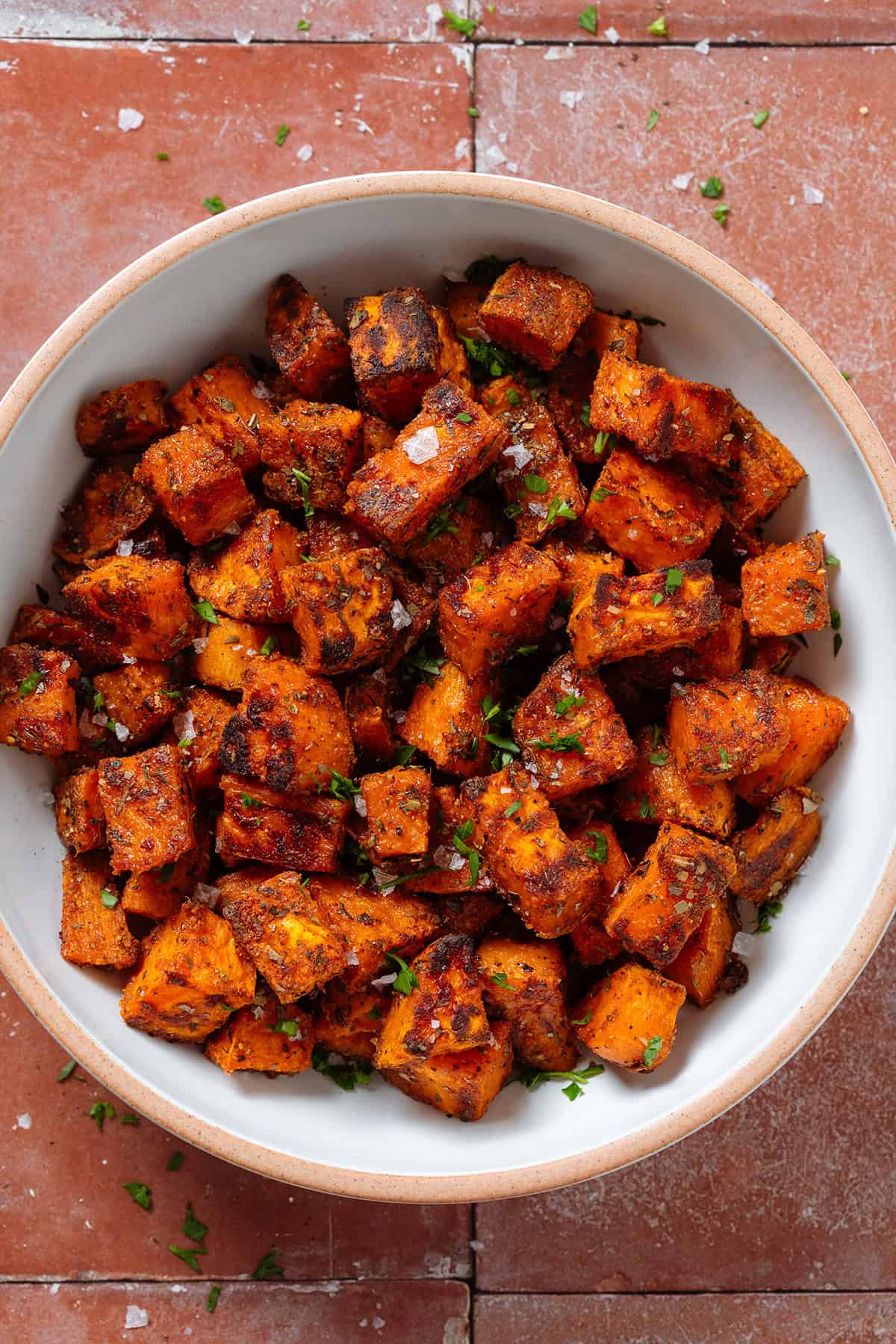 Crispy roasted diced sweet potatoes in a white bowl with a beige rim sprinkled with flaky sea salt and finely chopped fresh parsley.