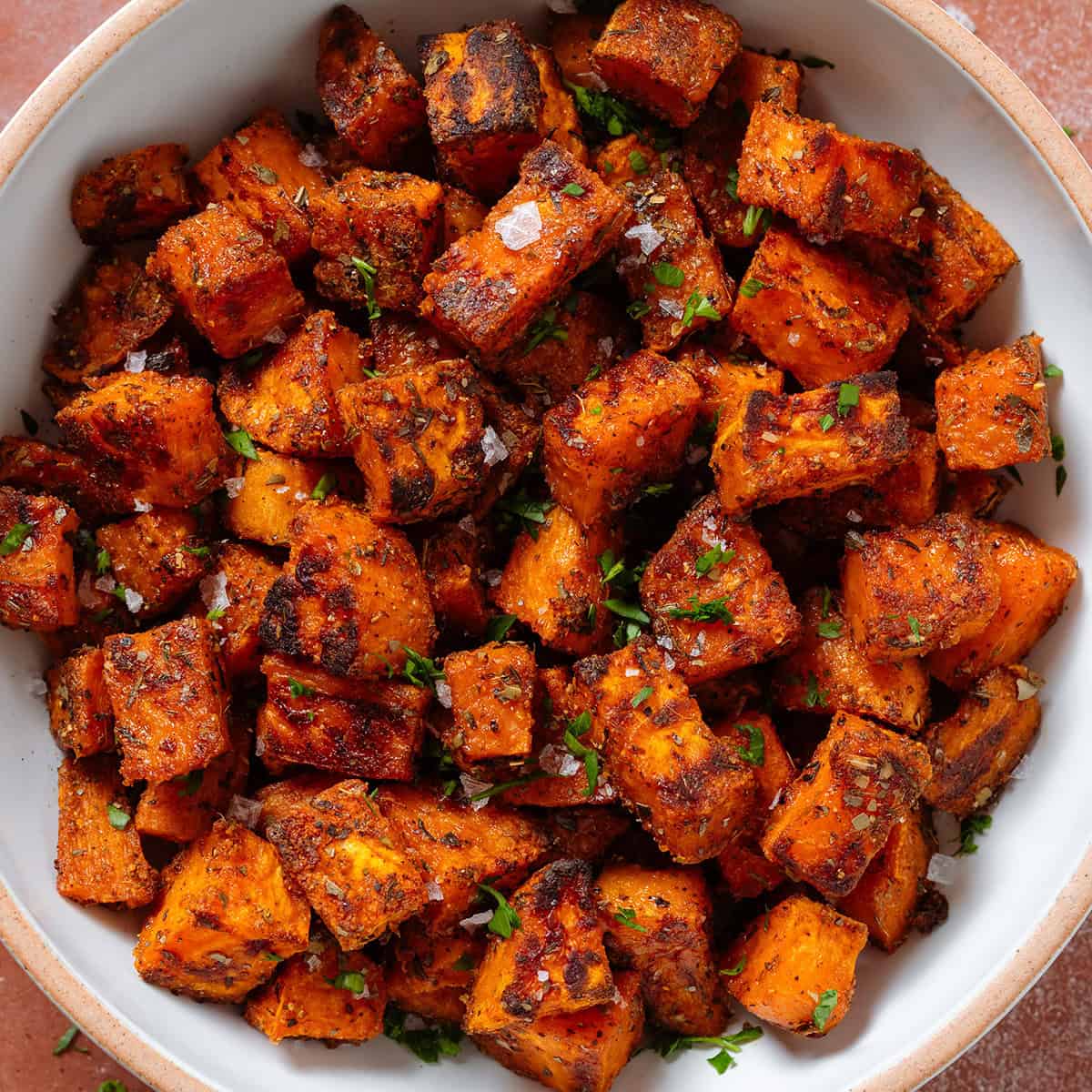 Crispy charred roasted diced sweet potatoes in a white bowl with a beige rim sprinkled with flaky sea salt and finely chopped fresh parsley.