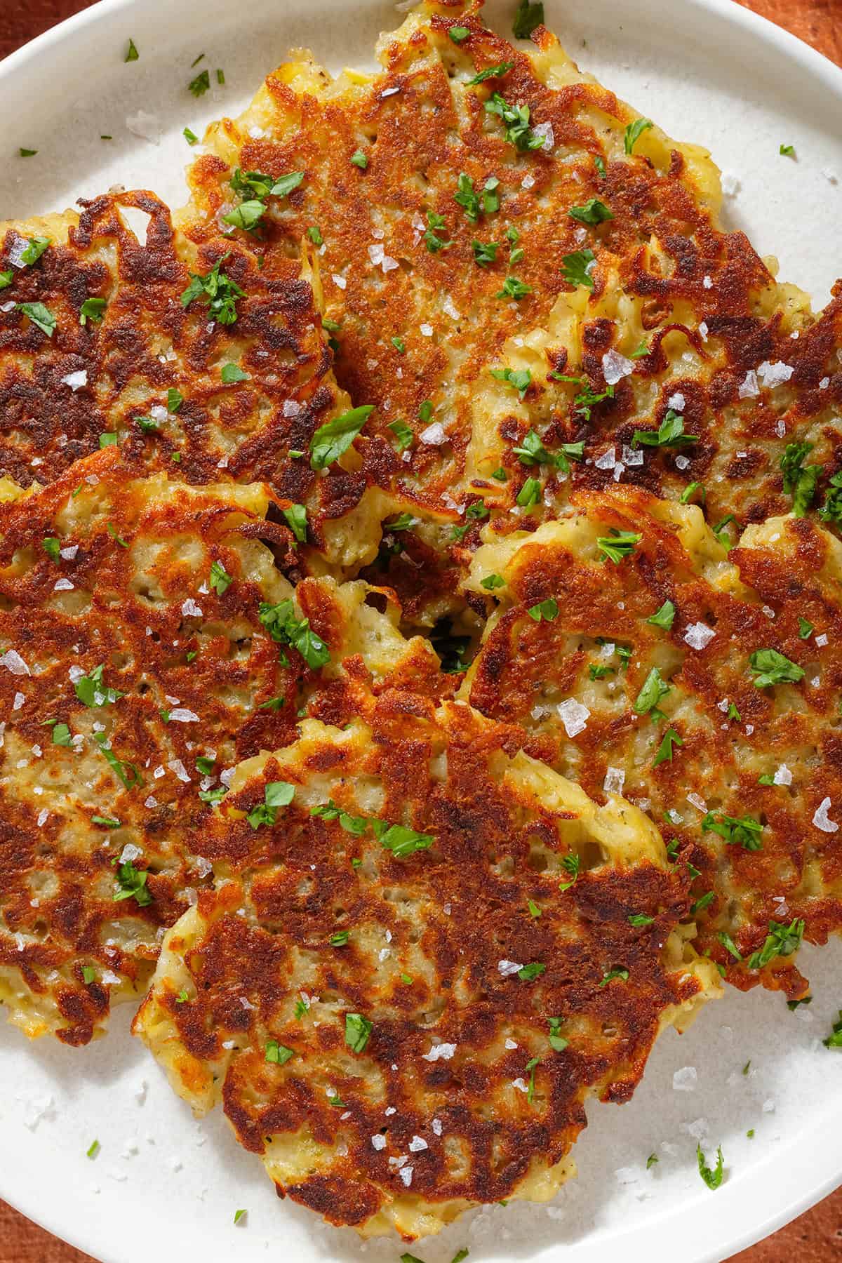 Golden brown potato fritters on a white plate sprinkled with finely chopped fresh parsley and flaky sea salt.