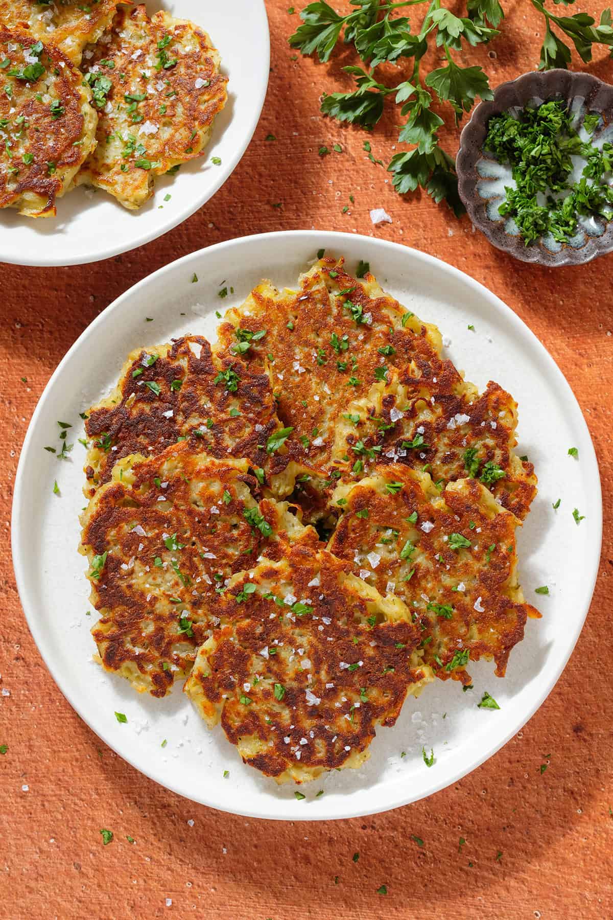 Golden brown potato fritters on a white plate sprinkled with finely chopped fresh parsley and flaky sea salt.