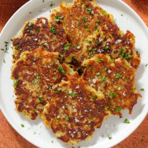 Golden brown potato fritters on a white plate sprinkled with finely chopped fresh parsley and flaky sea salt.