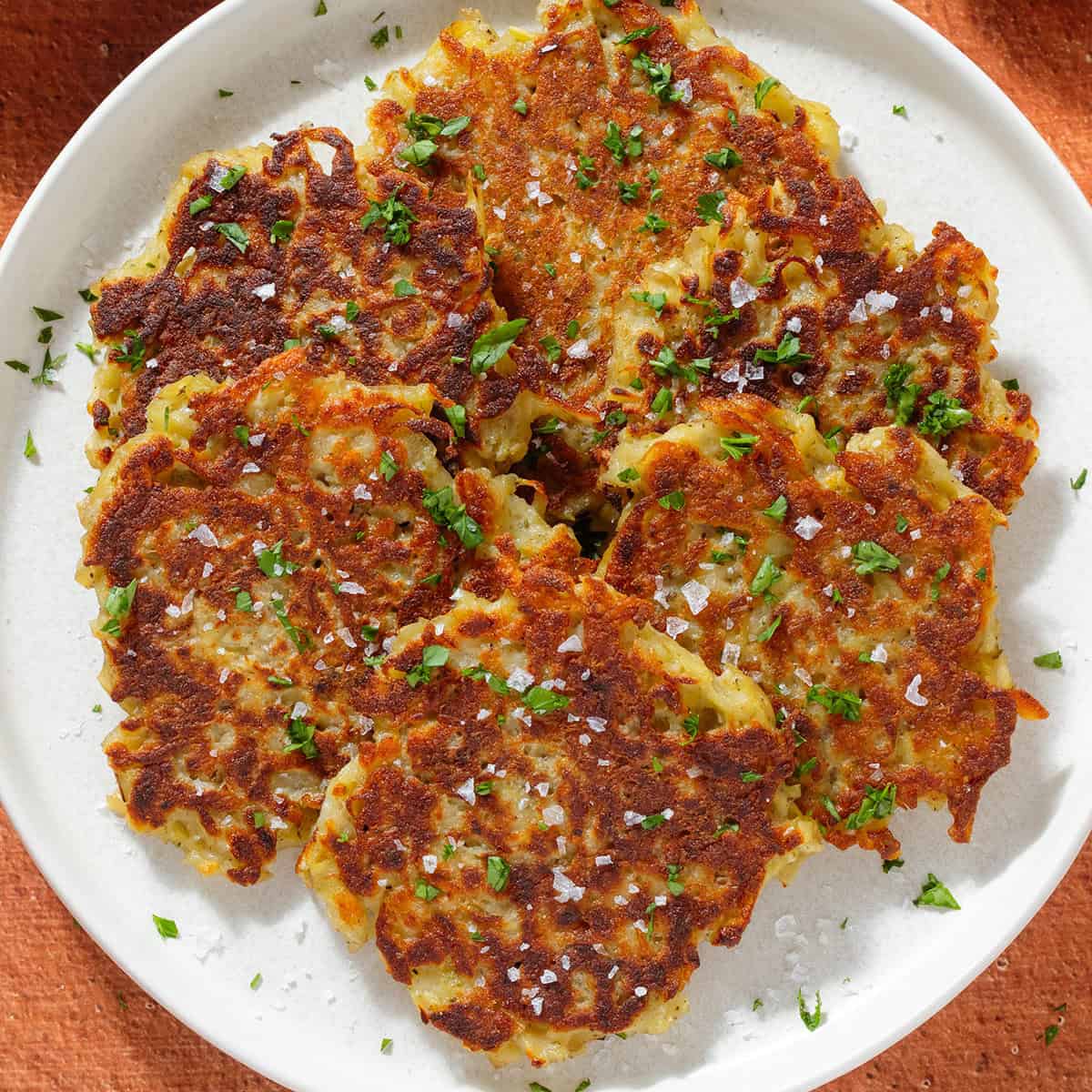 Golden brown potato fritters on a white plate sprinkled with finely chopped fresh parsley and flaky sea salt.