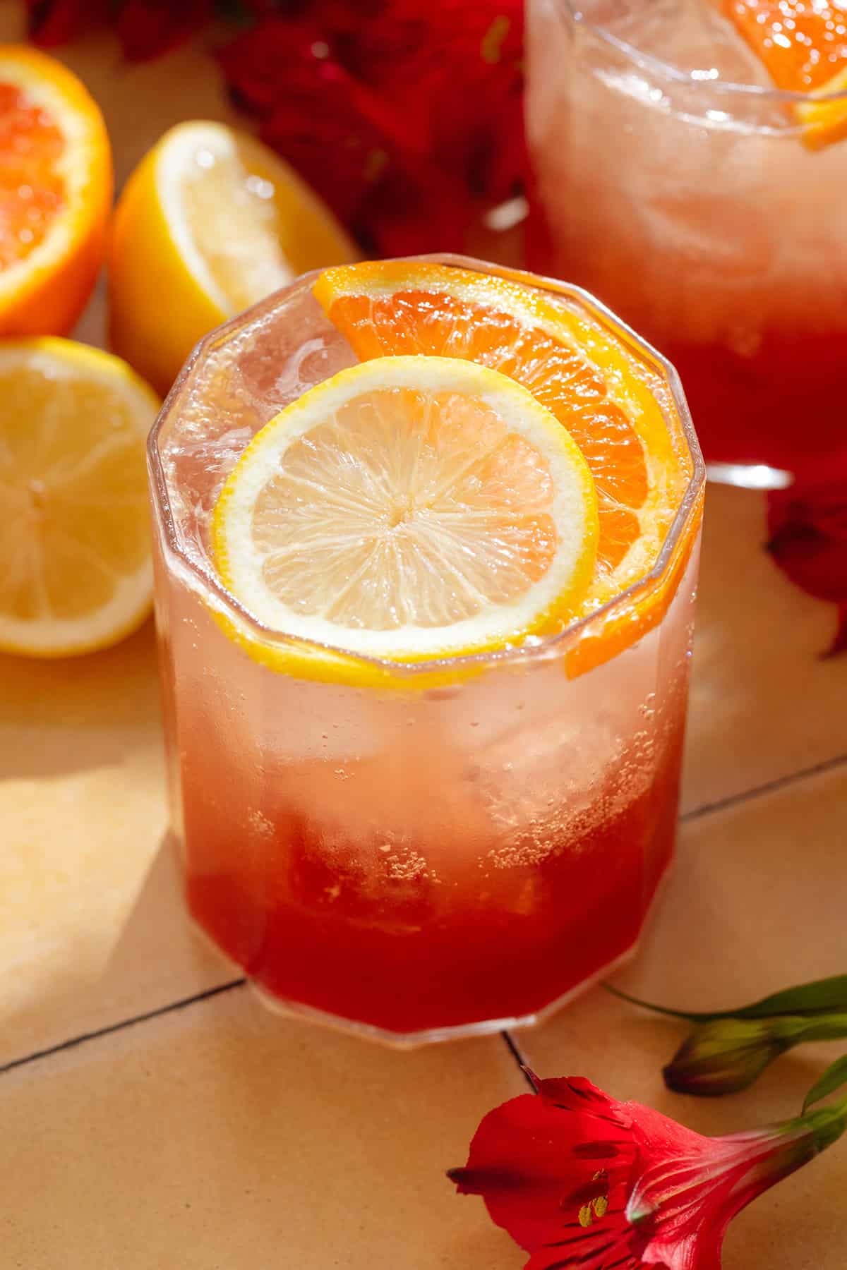A short glass with bright red hibiscus mocktail topped with sparkling water and garnished with a slice of orange and lemon.