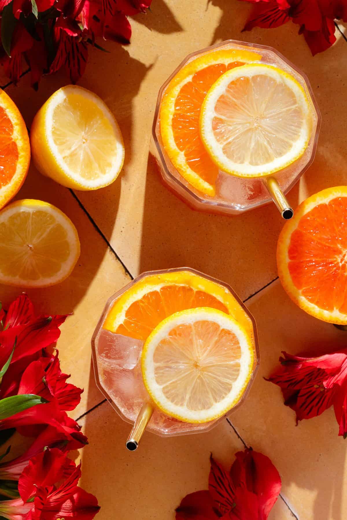 Two short glasses with bright red hibiscus mocktail topped with sparkling water and garnished with a slice of orange and lemon, with flower around the glasses.