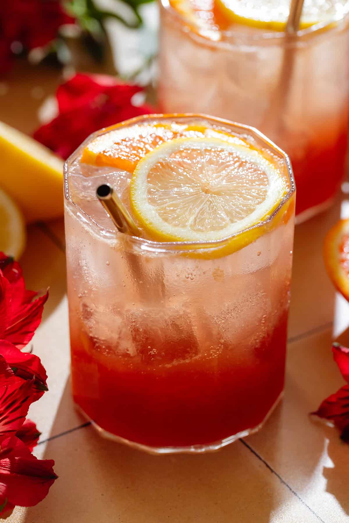 A short glass with bright red hibiscus mocktail topped with sparkling water and garnished with a slice of orange and lemon, with flower around the glass.