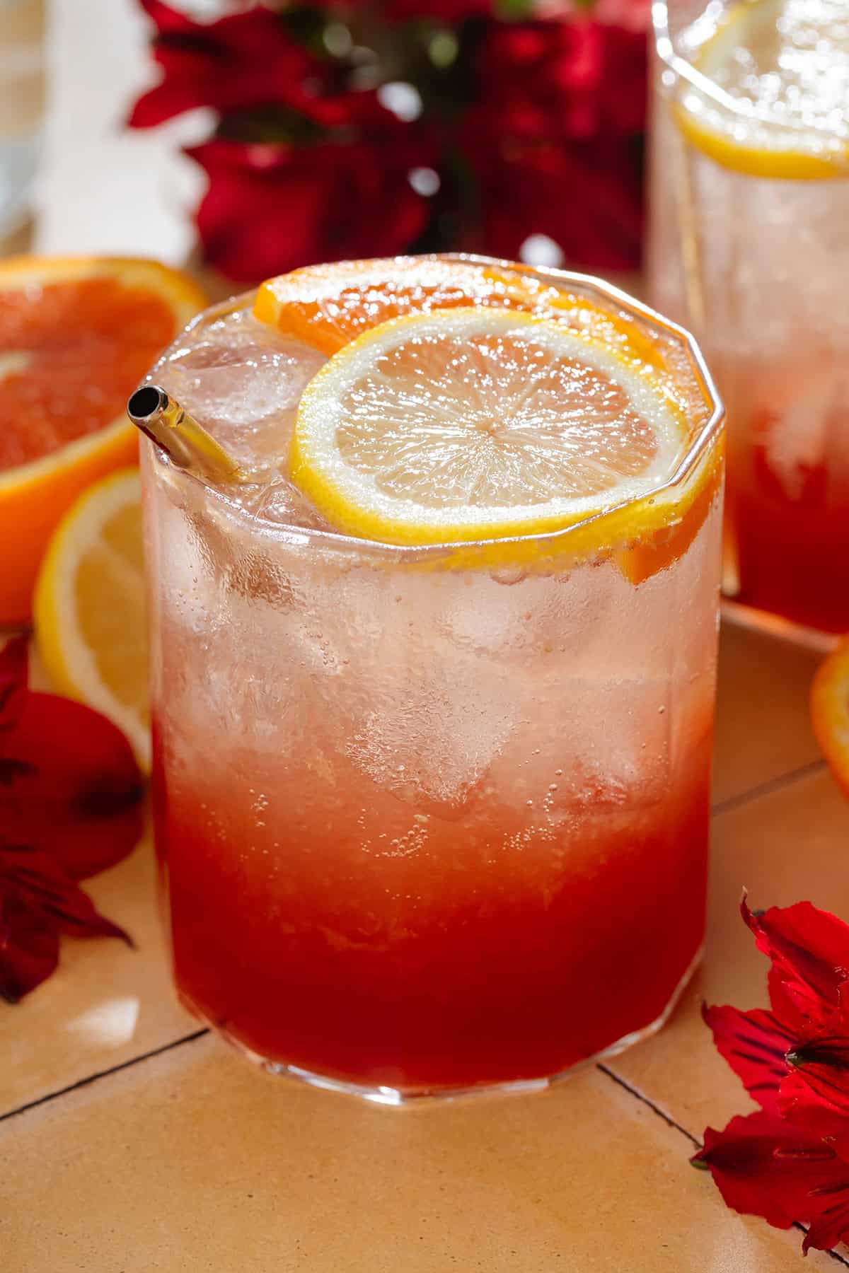 A short glass with bright red hibiscus mocktail topped with sparkling water and garnished with a slice of orange and lemon, with flower around the glass.