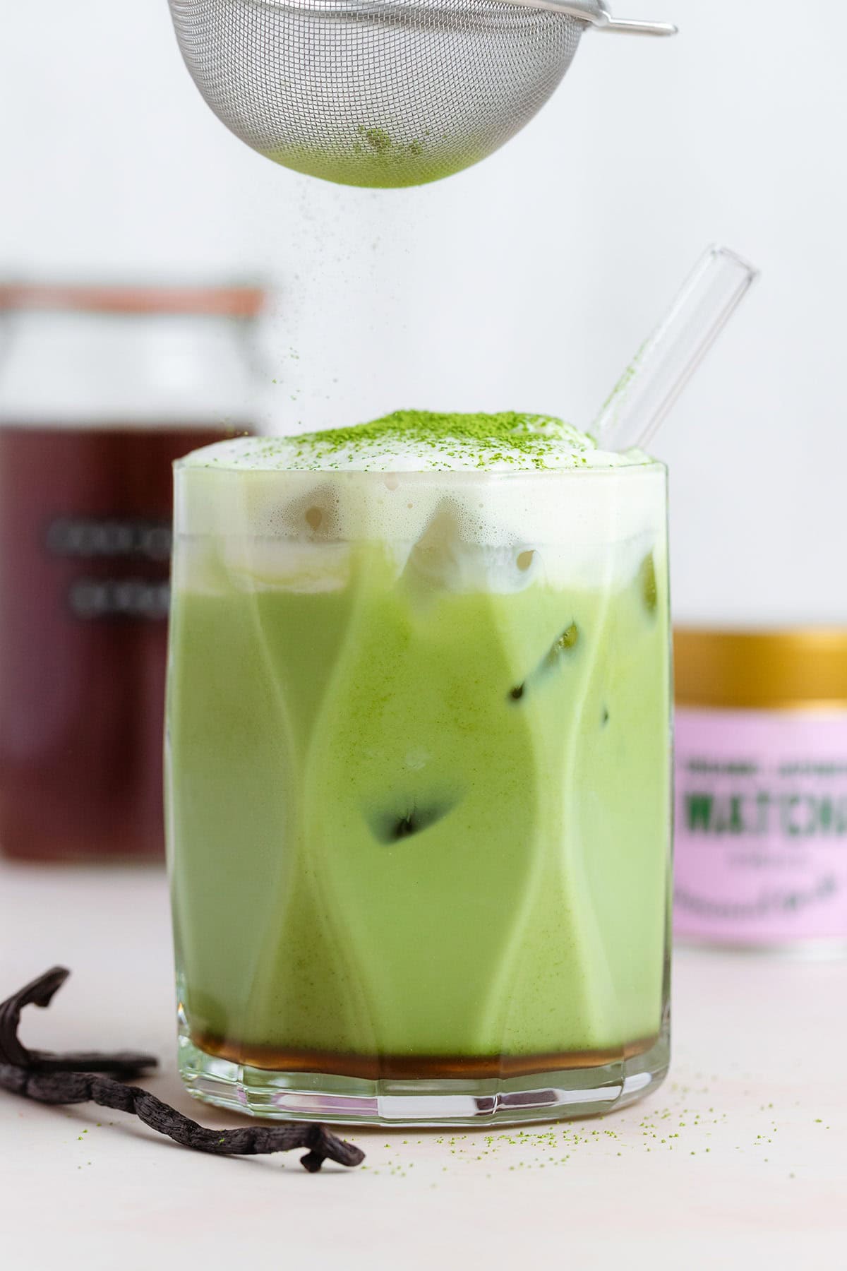 An iced matcha latte in a short glass being dusted with matcha powder for garnish.