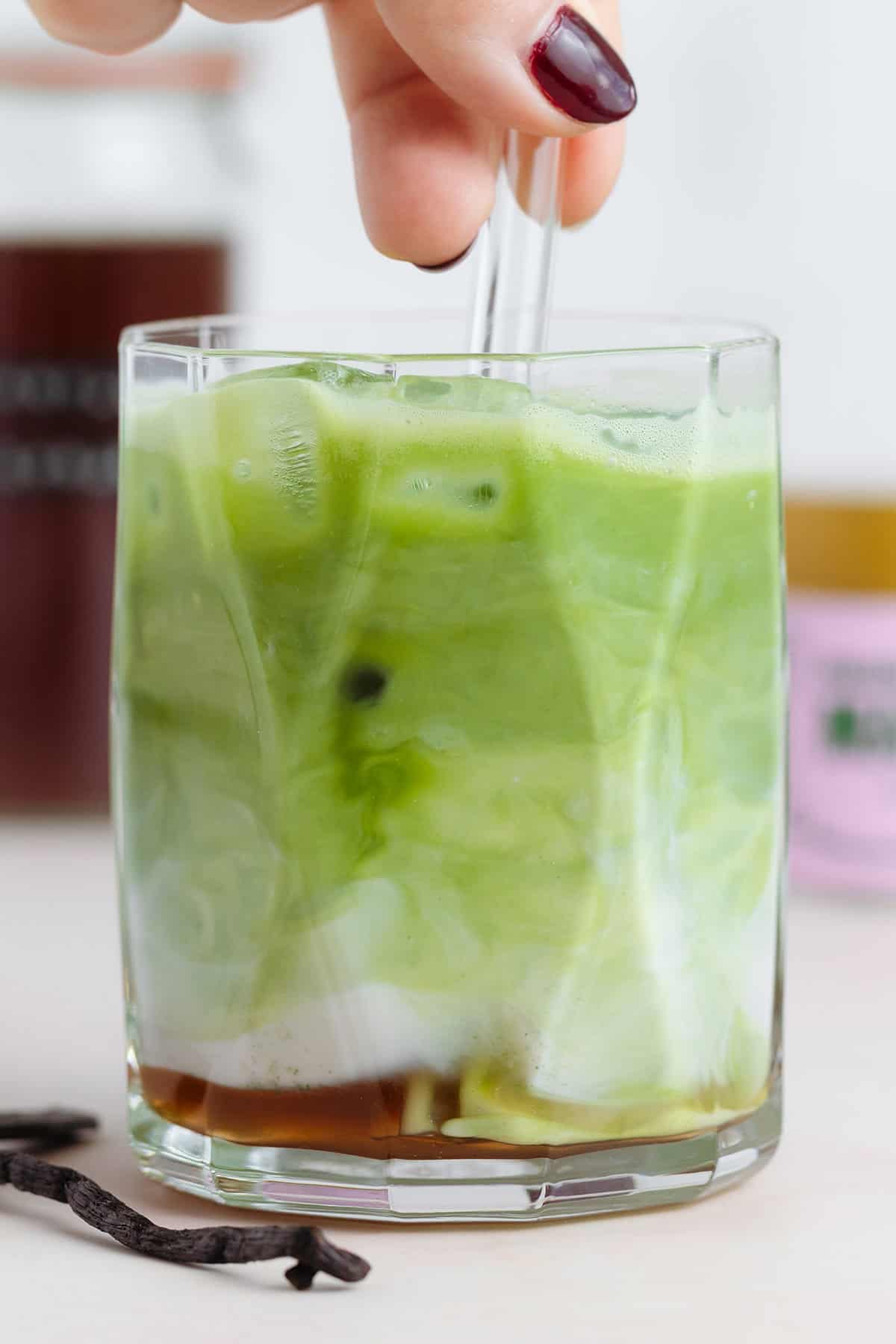 A hand stirring a matcha latte to mix in the syrup on the bottom of the glass.