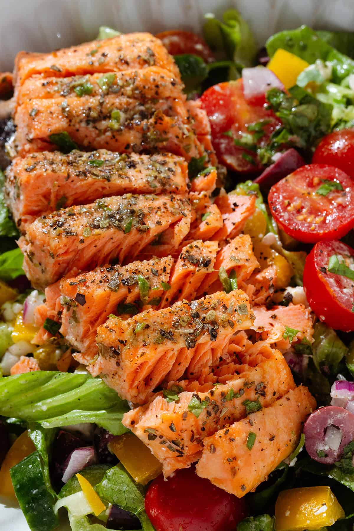 Colorful Greek salad in a white low bowl with a piece of roasted salmon sliced horizontally on top.