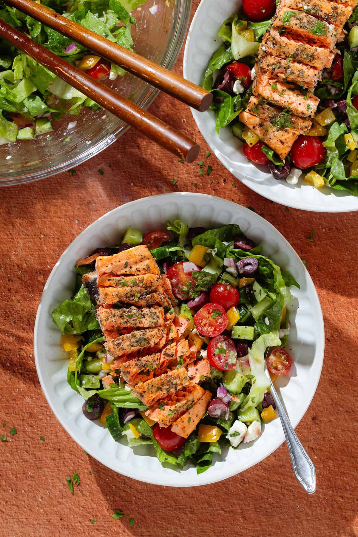 Colorful Greek salad in a white low bowls with a piece of roasted salmon sliced horizontally on top of each salad.