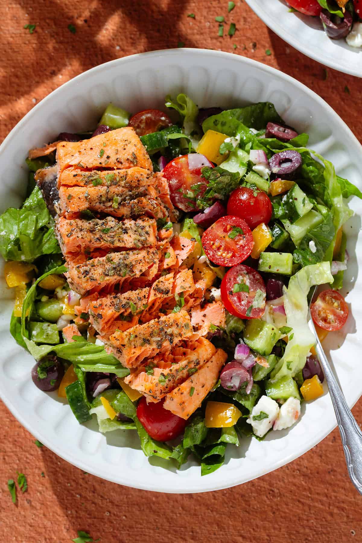 Colorful Greek salad in a white low bowl with a piece of roasted salmon sliced horizontally on top with a fork on the right.