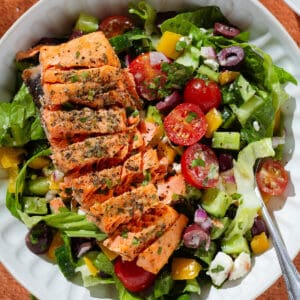 Colorful Greek salad in a white low bowl with a piece of roasted salmon sliced horizontally on top.