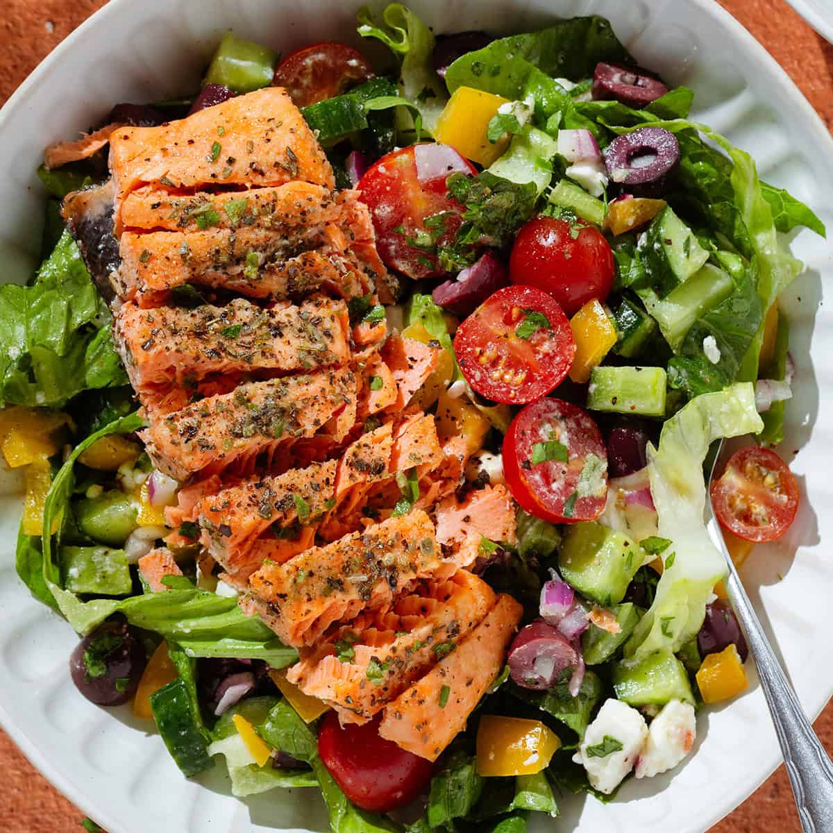 Colorful Greek salad in a white low bowl with a piece of roasted salmon sliced horizontally on top.