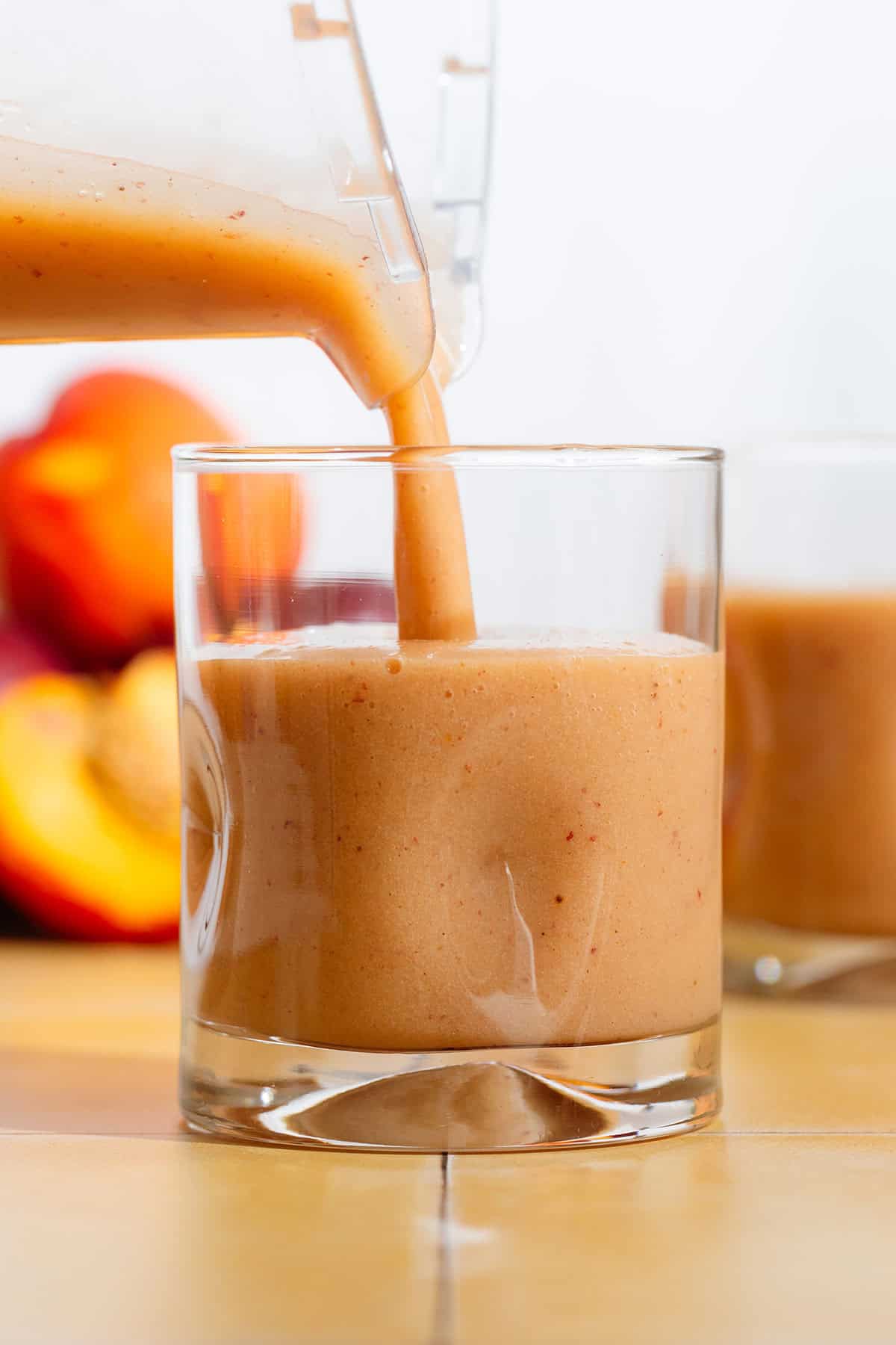 Nectarine smoothie being poured into a short glass on yellow tile table.