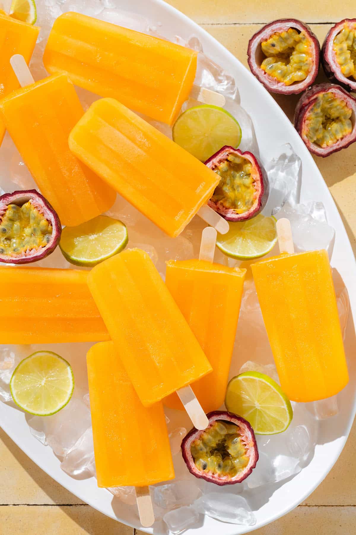 Bright orange popsicles on a white serving platter with ice, fresh passion fruit, and lime slices.