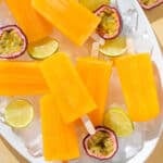 Bright orange popsicles on a white serving platter with ice, fresh passion fruit, and lime slices.