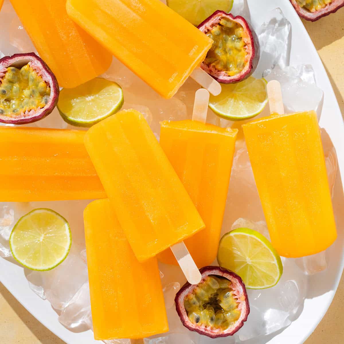 Bright orange popsicles on a white serving platter with ice, fresh passion fruit, and lime slices.