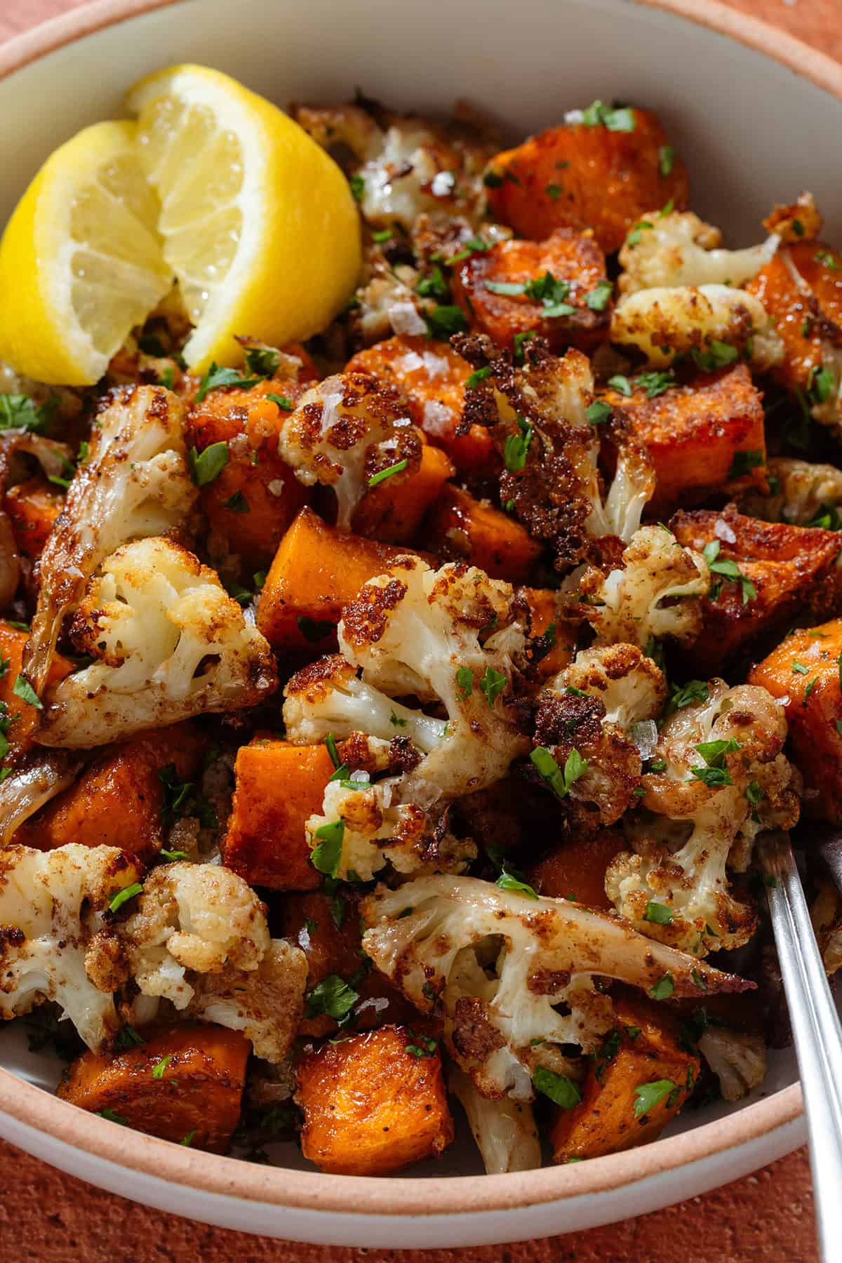 Roasted caramelized diced sweet potato and cauliflower florets in a white bowl garnished with flaky sea salt and chopped fresh parsley and with two lemon wedges on the left and a fork on the right.
