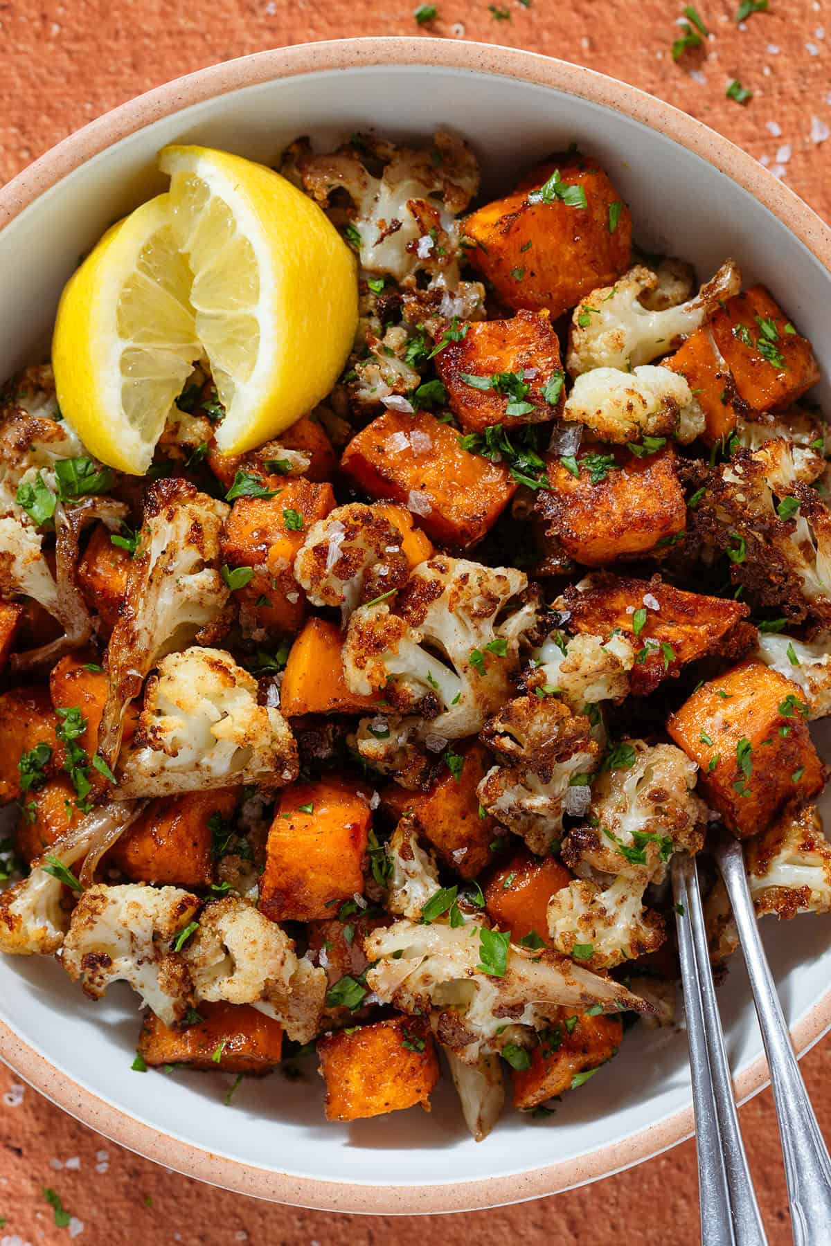 Roasted caramelized diced sweet potato and cauliflower florets in a white bowl sprinkled with flaky sea salt, chopped fresh parsley, and two lemon wedges on the left, with two forks on the right.