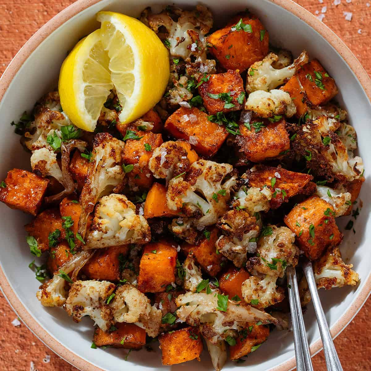 Roasted caramelized diced sweet potato and cauliflower florets in a white bowl sprinkled with flaky sea salt and chopped fresh parsley and with two lemon wedges on the left.