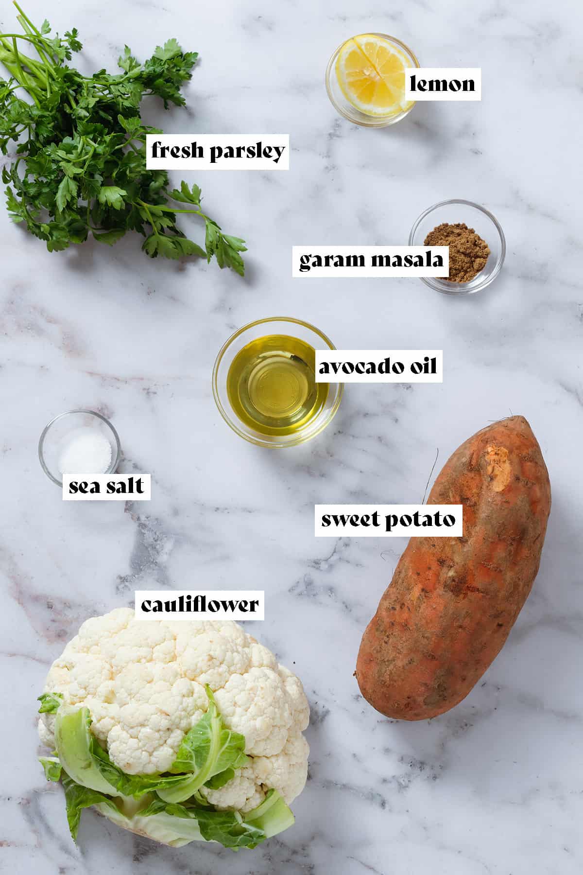One sweet potato, a head of cauliflower, fresh parsley, lemon, avocado oil, sea salt, and garam masala in small bowls laid out on a marble background.