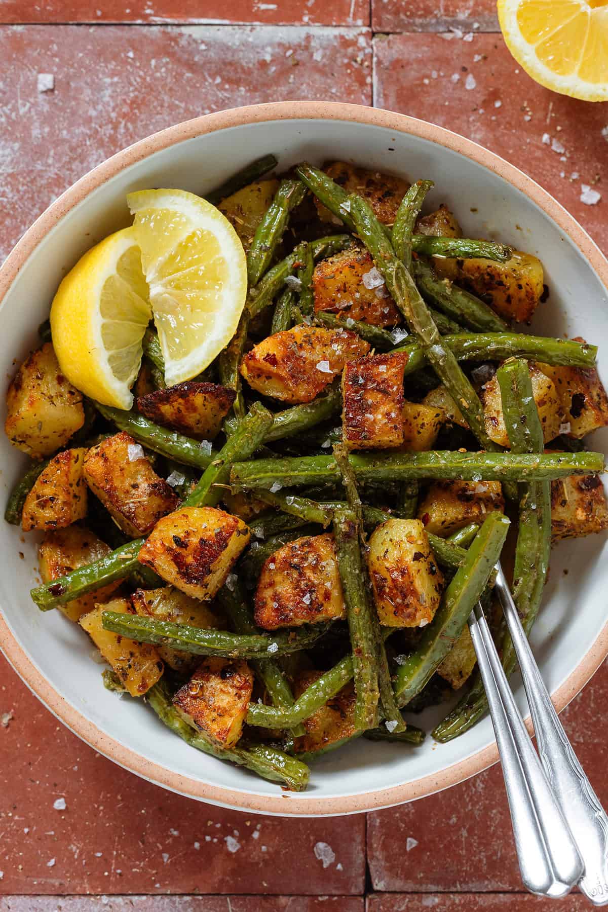 Crispy golden roasted diced potatoes and green beans in a white bowl with two lemon wedges on the left and two forks inserted on the right.