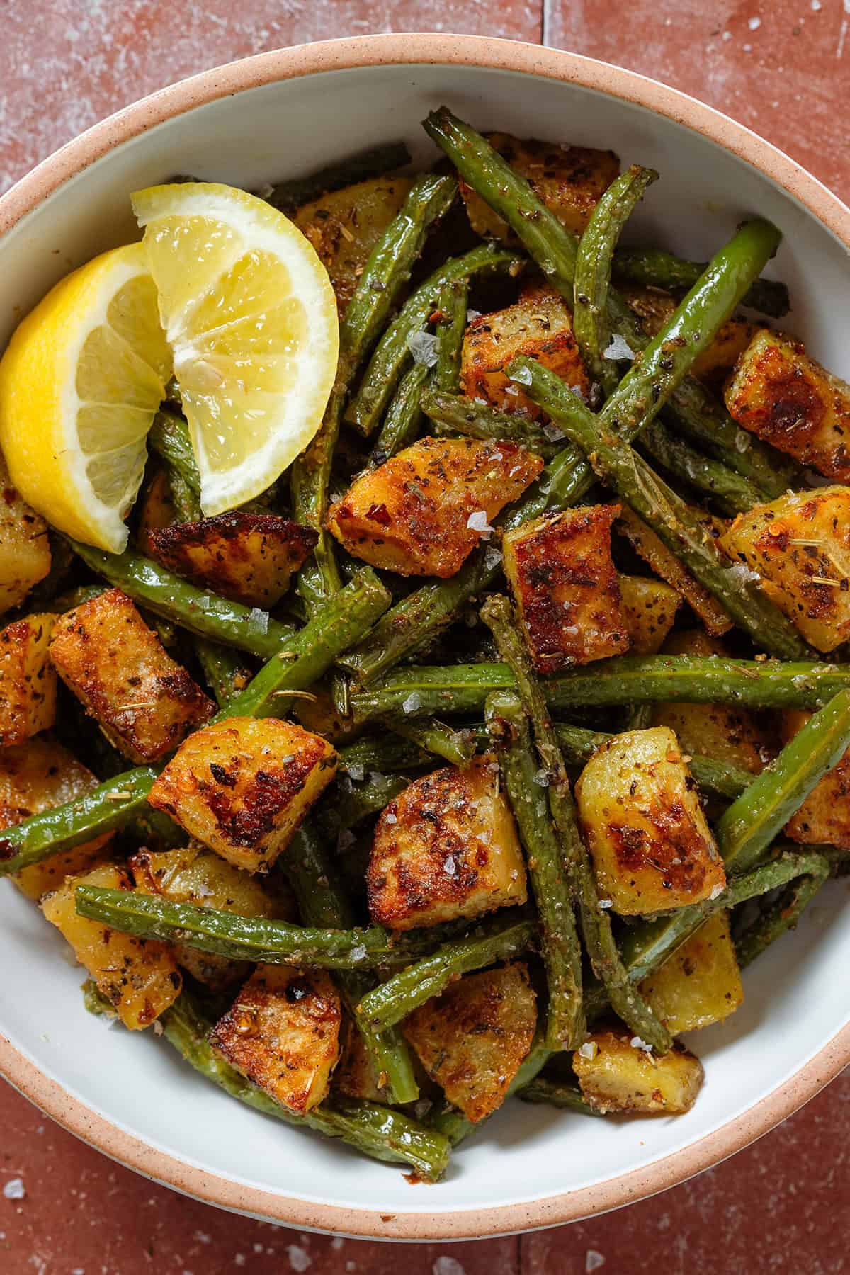 Crispy golden roasted diced potatoes and green beans in a white bowl with two lemon wedges on the left.