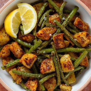 Crispy golden roasted diced potatoes and green beans in a white bowl with two lemon wedges on the left.