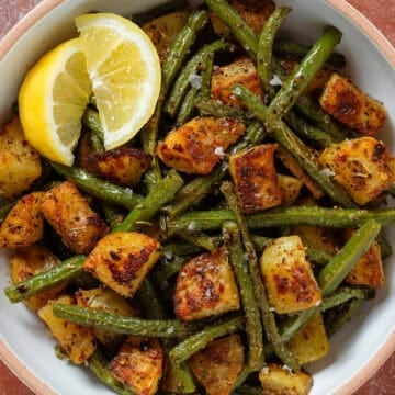Crispy golden roasted diced potatoes and green beans in a white bowl with two lemon wedges on the left.