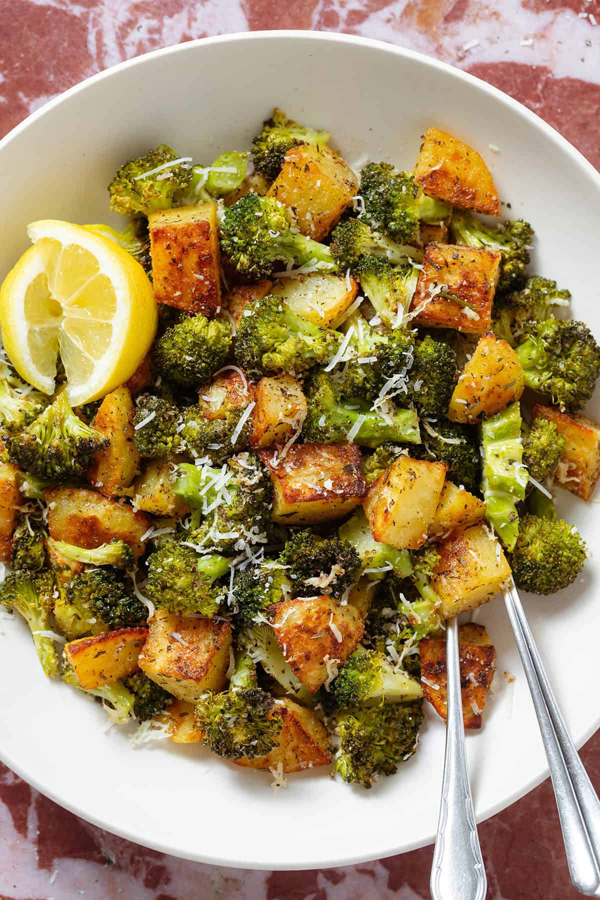 Roasted broccoli florets and crispy diced potatoes sprinkled with grated parmesan in a white bowl garnished with two lemon wedges on the left.