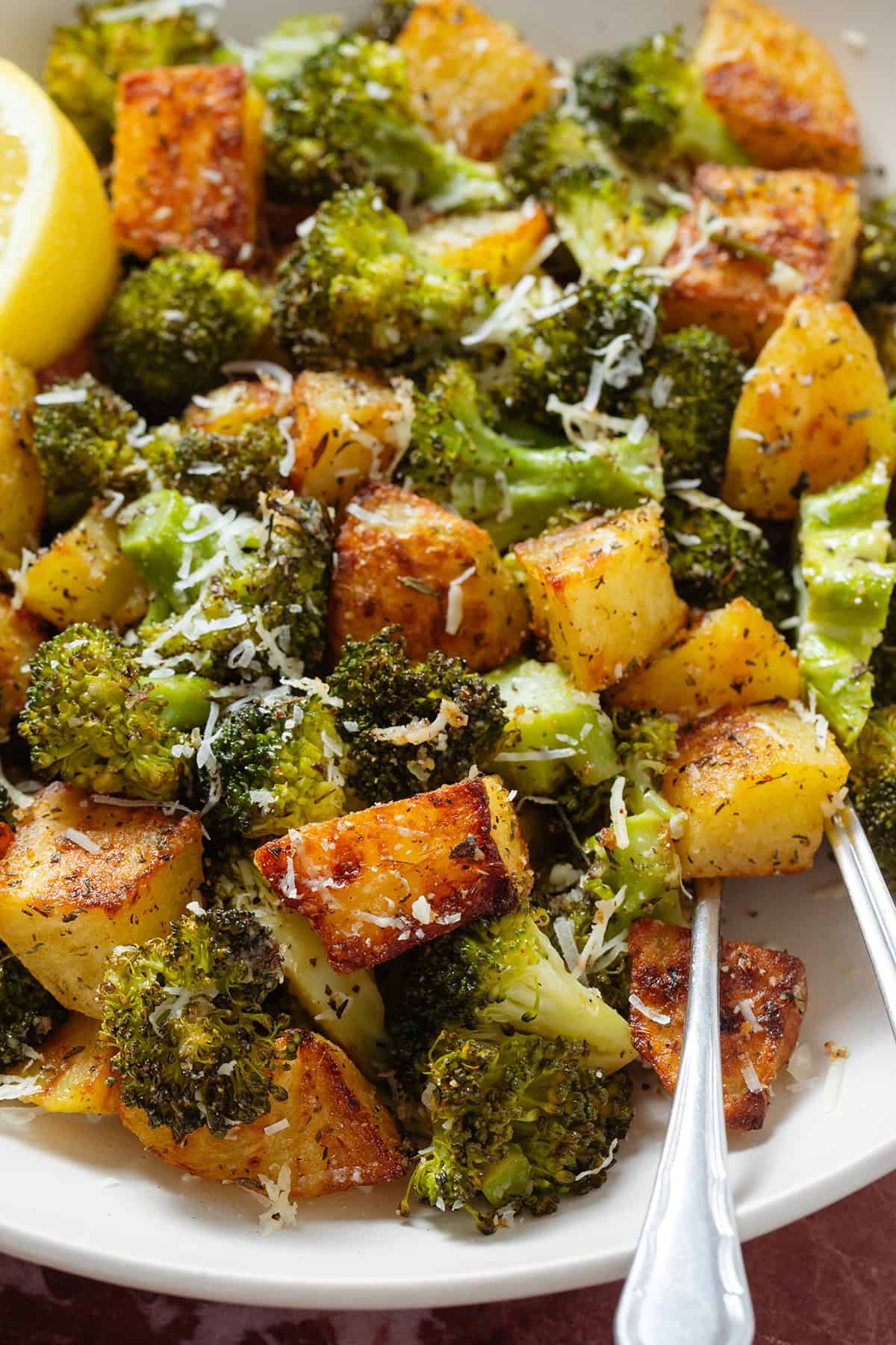 Roasted broccoli florets and crispy diced potatoes sprinkled with grated parmesan in a white bowl with two forks on the right.