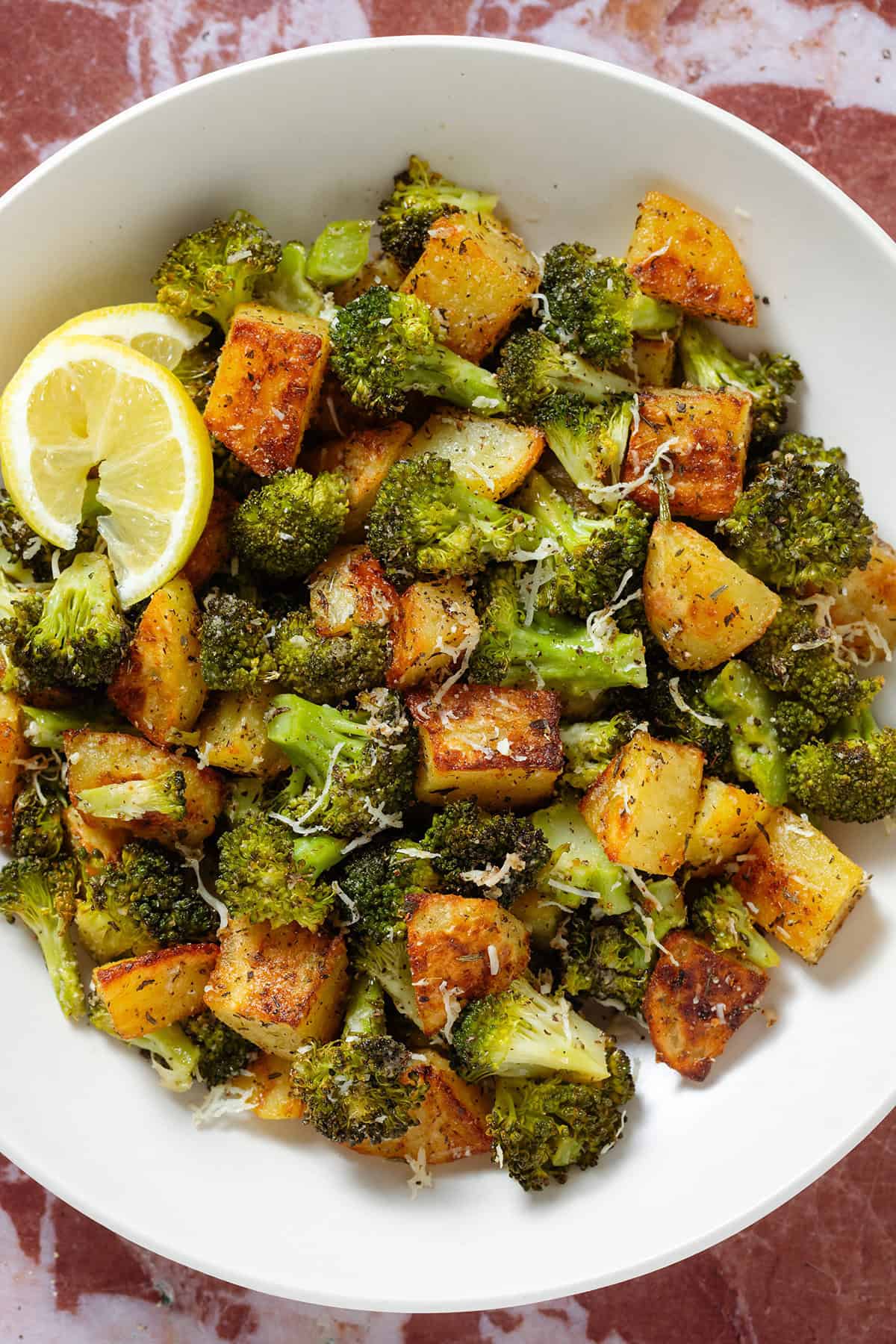 Roasted broccoli florets and crispy diced potatoes sprinkled with grated parmesan in a white bowl garnished with two lemon wedges on the left.