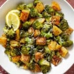 Roasted broccoli florets and crispy diced potatoes sprinkled with grated parmesan in a white bowl garnished with two lemon wedges on the left.