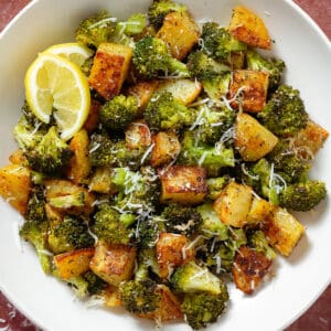Roasted broccoli florets and crispy diced potatoes sprinkled with grated parmesan in a white bowl garnished with two lemon wedges on the left.