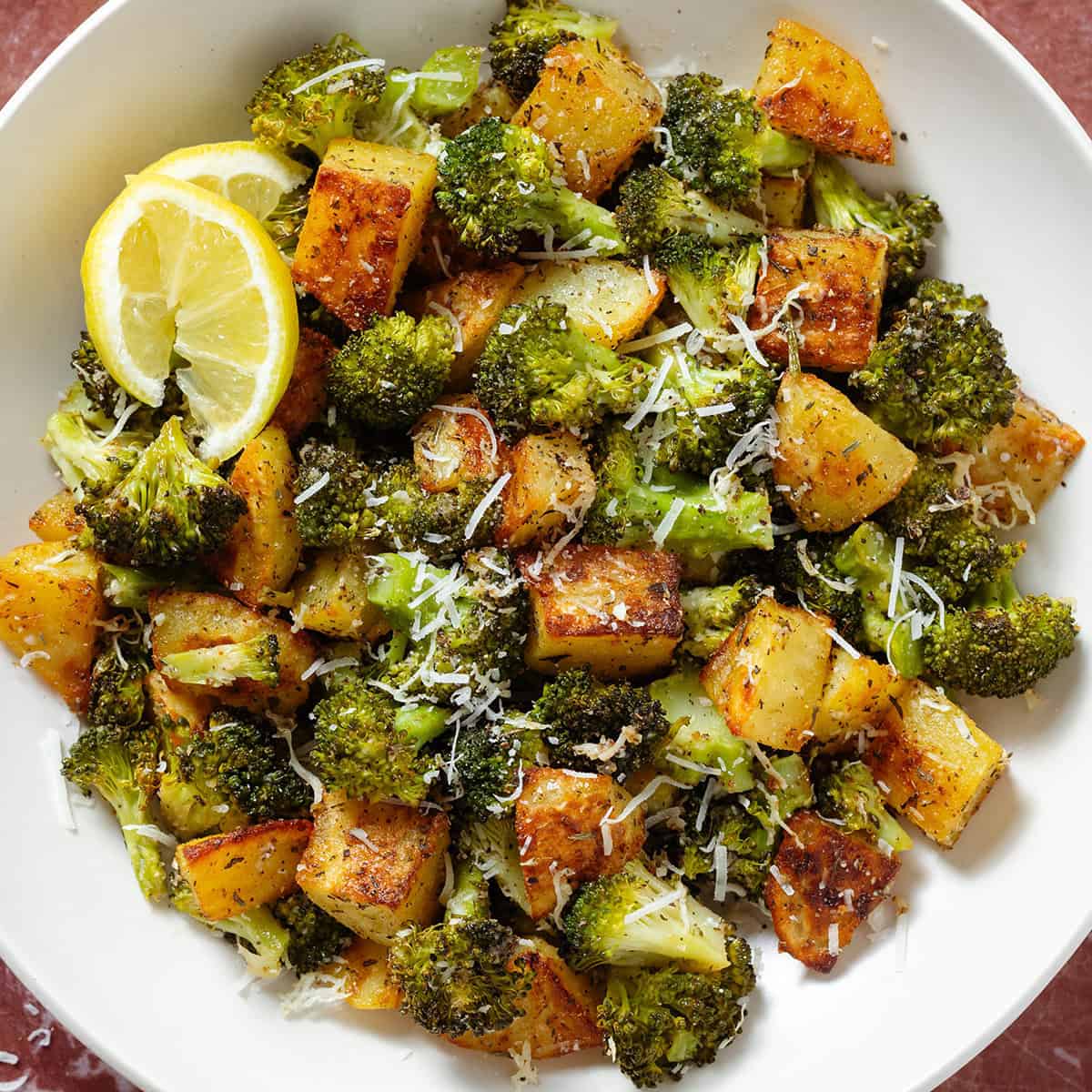 Roasted broccoli florets and crispy diced potatoes sprinkled with grated parmesan in a white bowl garnished with two lemon wedges on the left.