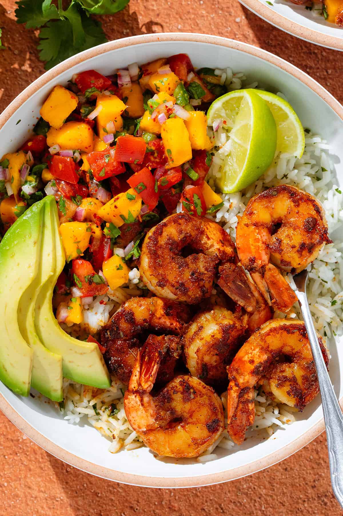 A white bowl with cilantro rice, spicy shrimp, mango pico de gallo, slice avocado, and two lemon wedges.