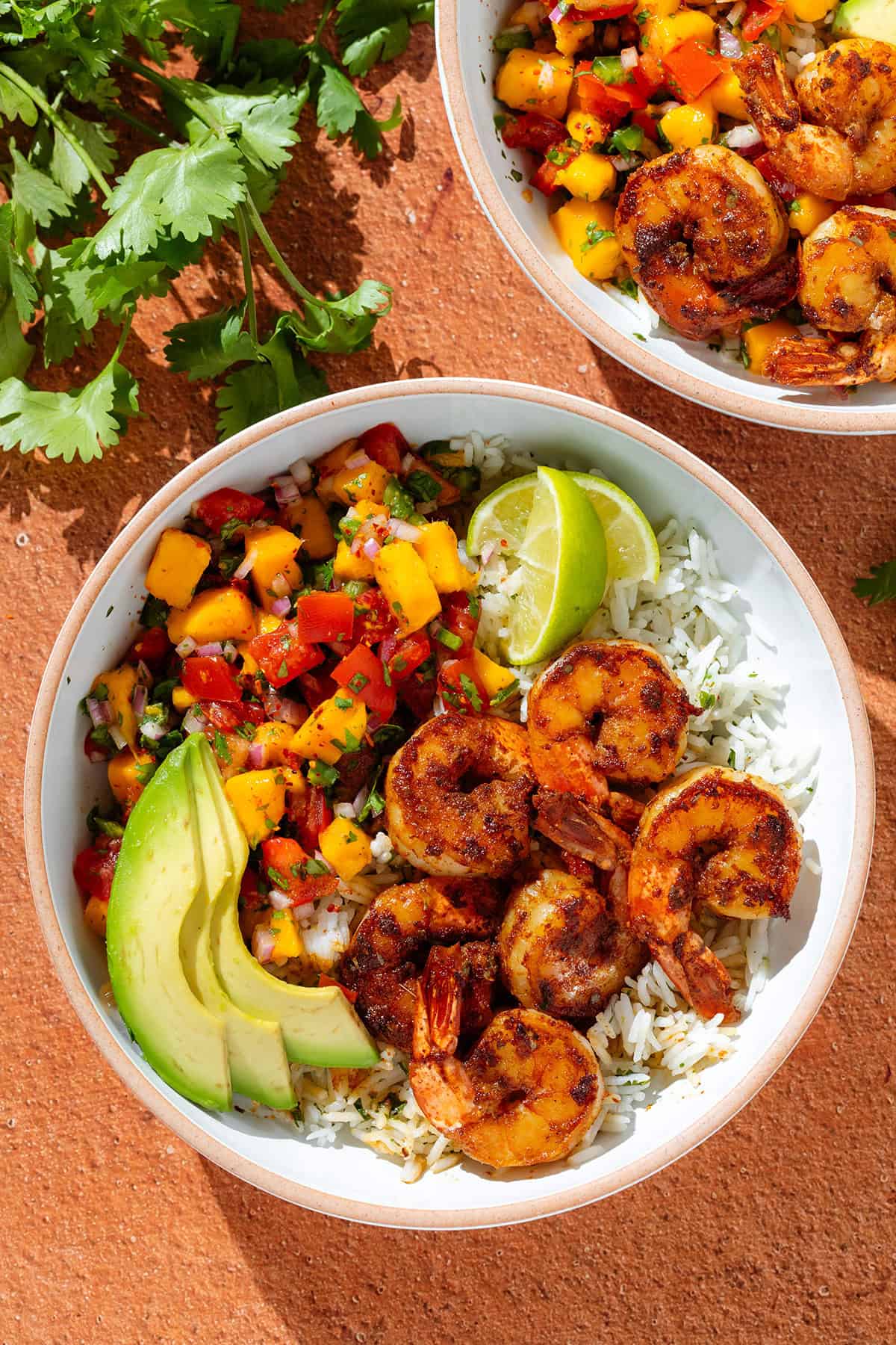 Two white bowls with cilantro rice, spicy shrimp, mango pico de gallo, slice avocado, and two lemon wedges.