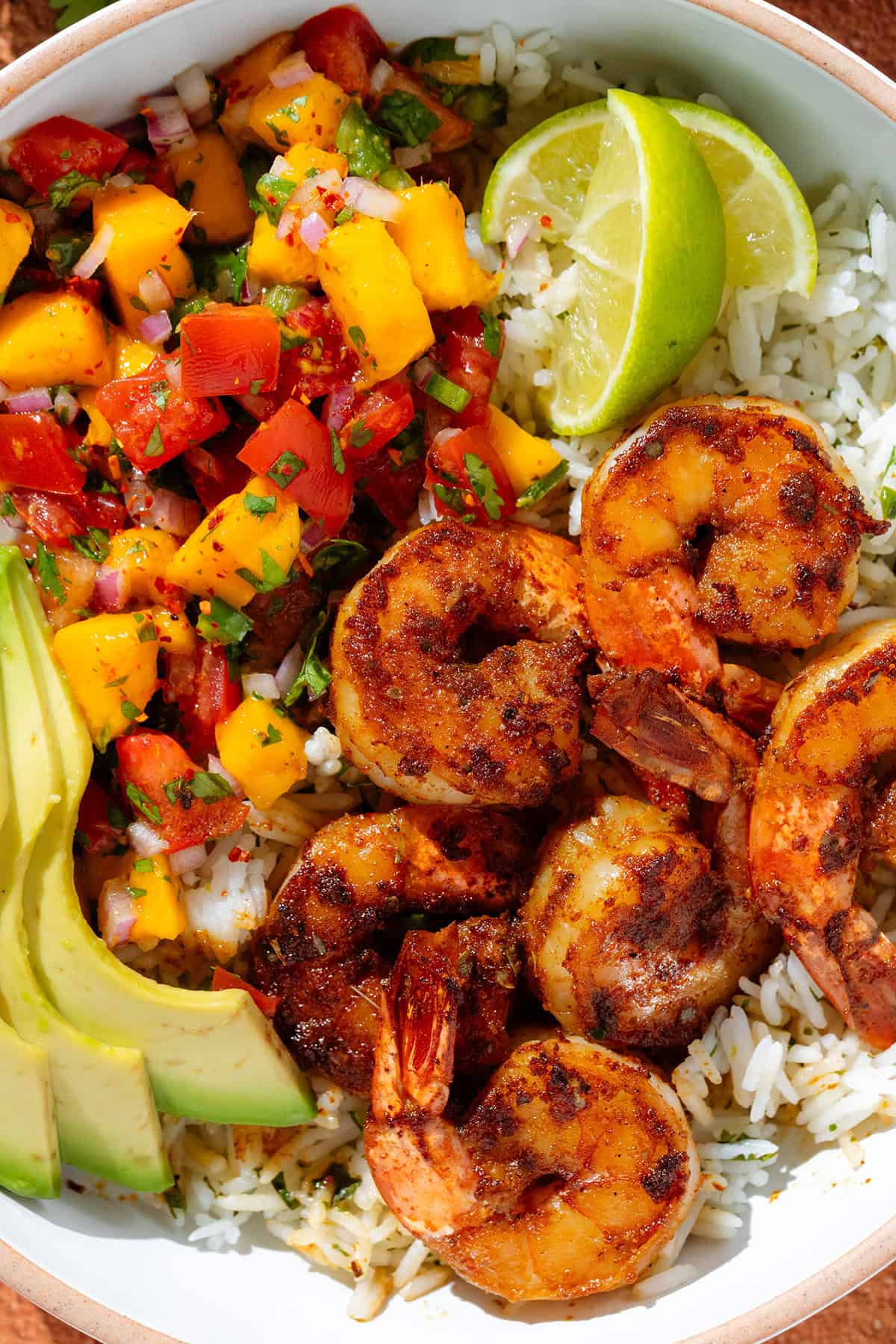 A bowl with cilantro rice, spicy shrimp, mango pico de gallo, slice avocado, and two lemon wedges.