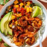 A bowl with cilantro rice, spicy shrimp, mango pico de gallo, slice avocado, and two lemon wedges.