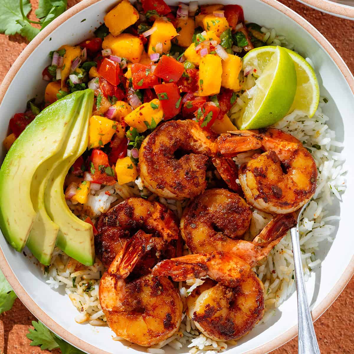 A bowl with cilantro rice, spicy shrimp, mango pico de gallo, slice avocado, and two lemon wedges.