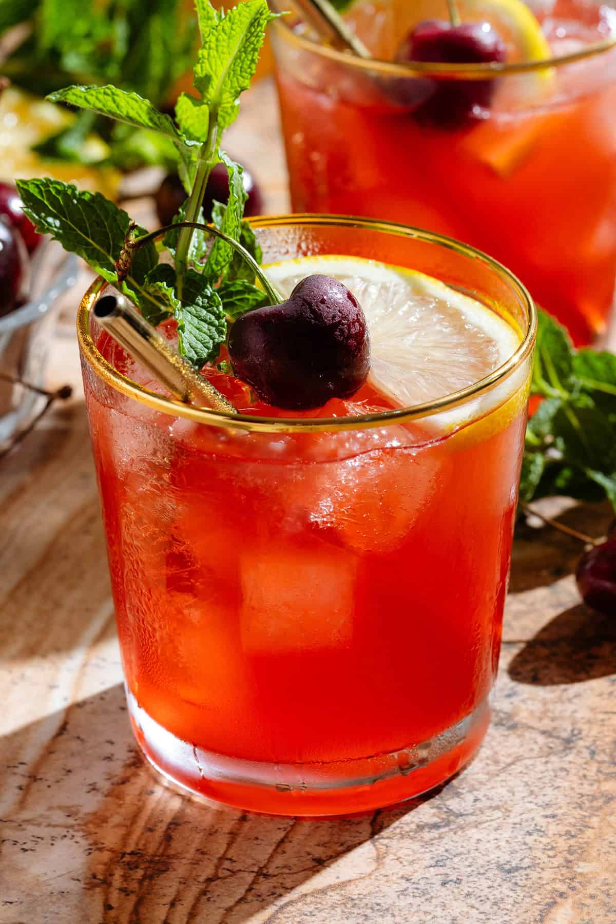 A red bourbon cherry cocktail in a short glass with a gold rim garnished with a slice of lemon, a fresh cherry, and a sprig of fresh mint.